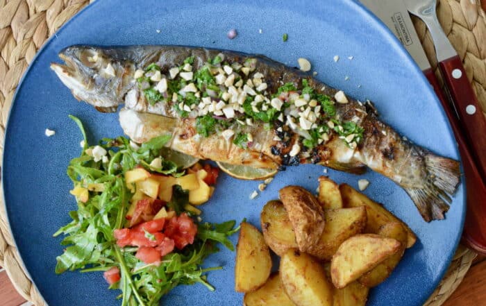 Gegrillte Forelle auf blauem Teller mit gerösteten Kartoffeln und Ruccolasalat mit Tomatenstückchen