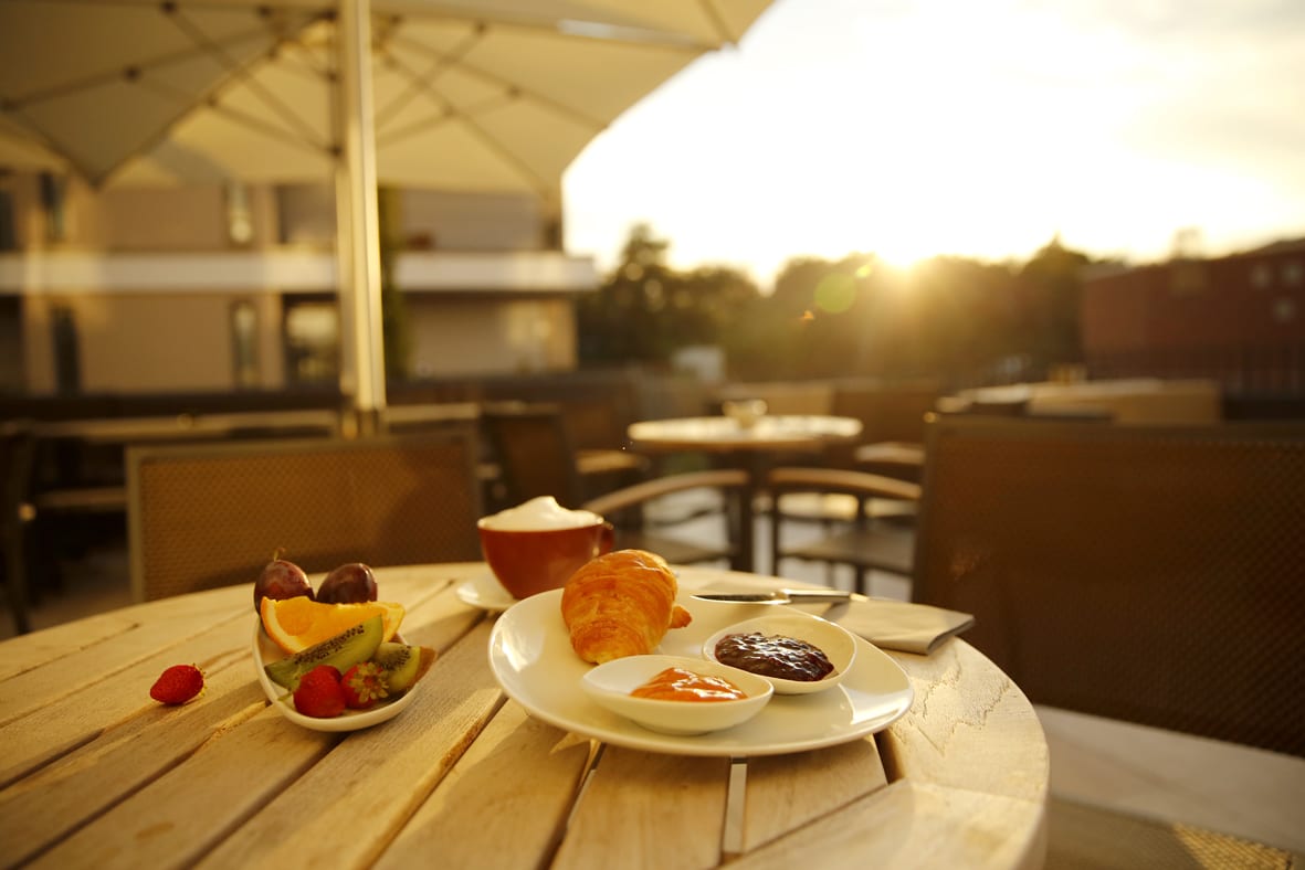 Hotel Landau Frühstück Frühstück mit Croissant, Marmelade Schale mit frischem Ost und einer Tasse Cappuccino auf einem Holttisch auf der Terrase des Boutique-Hotels Maximilians in Landau in der Pfalz im Sonnenschein. Im Hintergrund ein aufgespannter Sonnenschirm.