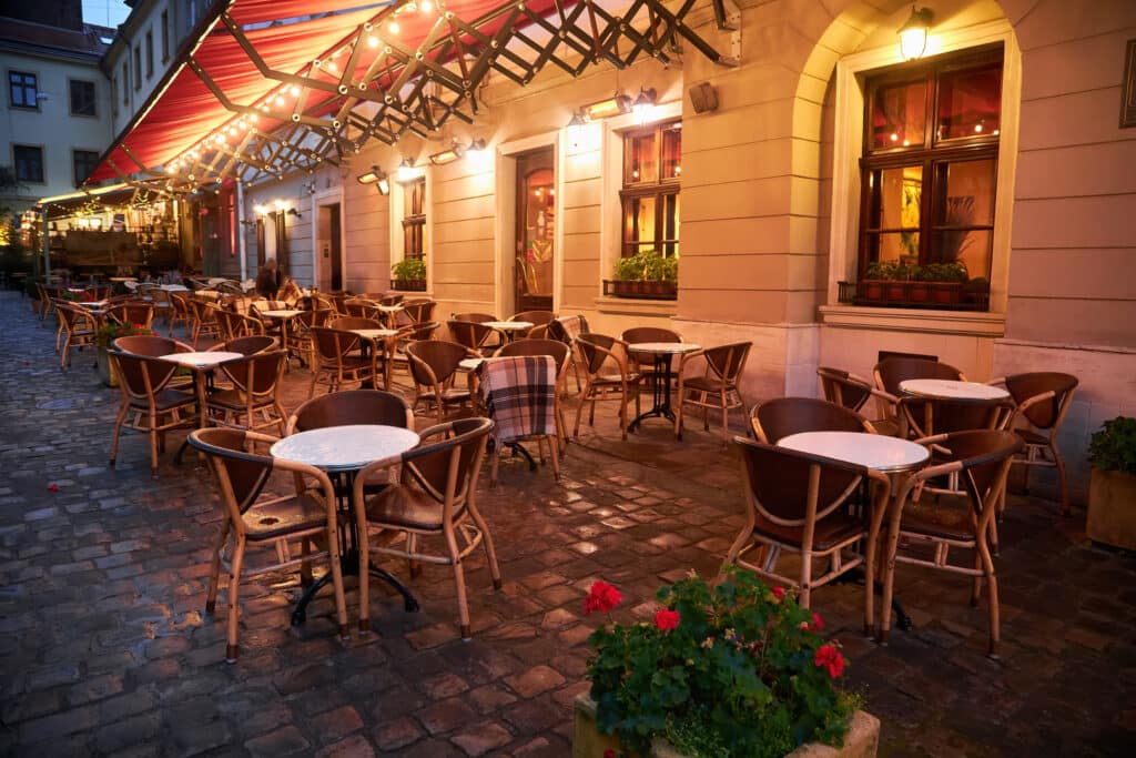 tables and chairs of a street restaurant in evening Beleuchtete Restaurantaußenbereich mit runden Tischen und Korbstühlen im Dämmerlicht