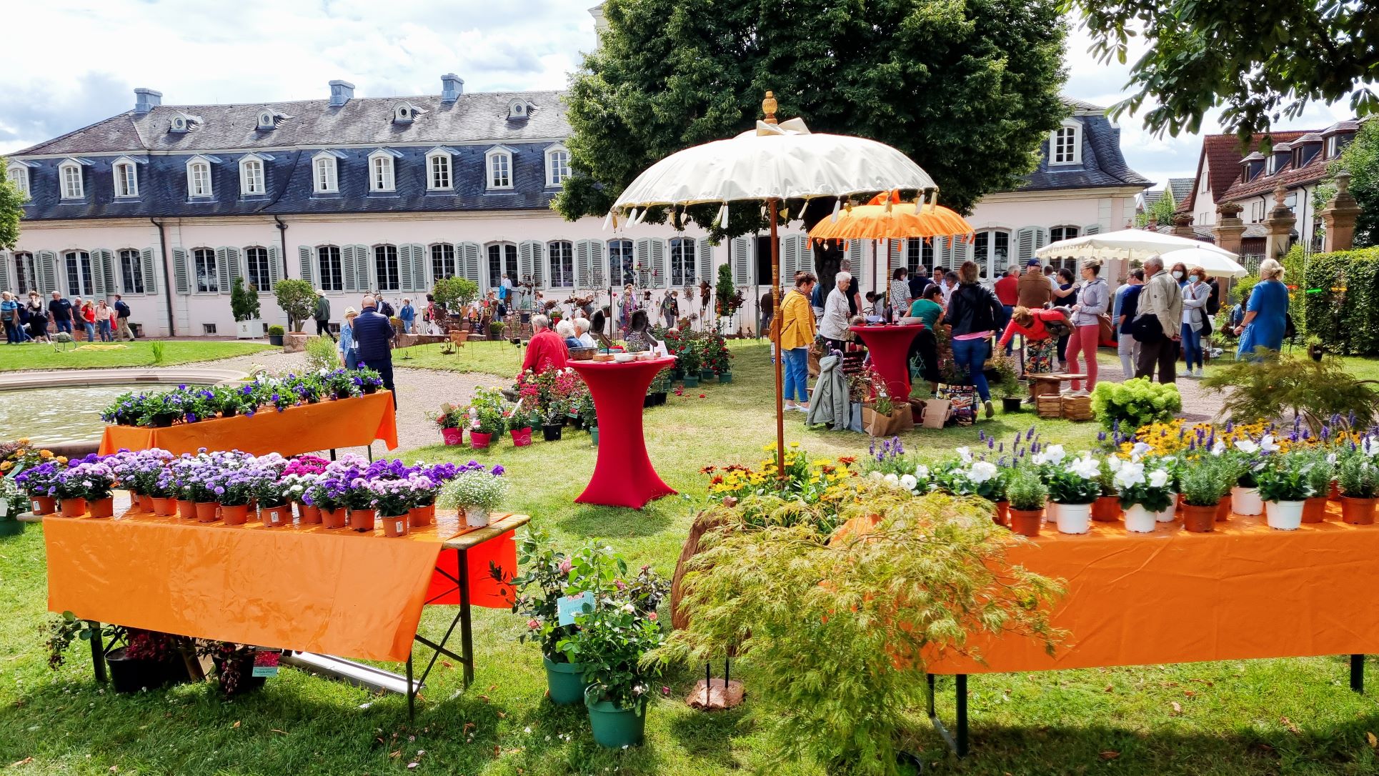 Genuss- und Gartenfest auf Schloss Braunshardt - Menschen schlendern durch den Schlosspark mit Ausstellern und Blumen, Sonnenschirmen und Stehtischen mit roten Hussen.