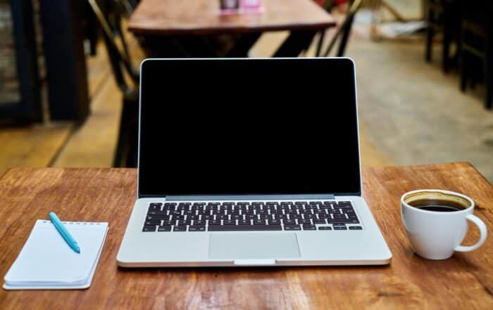 kaffeetase ohne Untertasse neben Laptop und Notitzblock mit hellblauem Kugelschreiber auf einem Holztisch in einem Restaurant. Der Hintergrund ist unscharf der Screen ist schwarz.