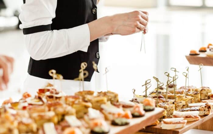 Service Mitarbeiterin in weißer Bluse und schwarzer Weste steht hinter Catering Tabletts mit kleinen, Häppchen