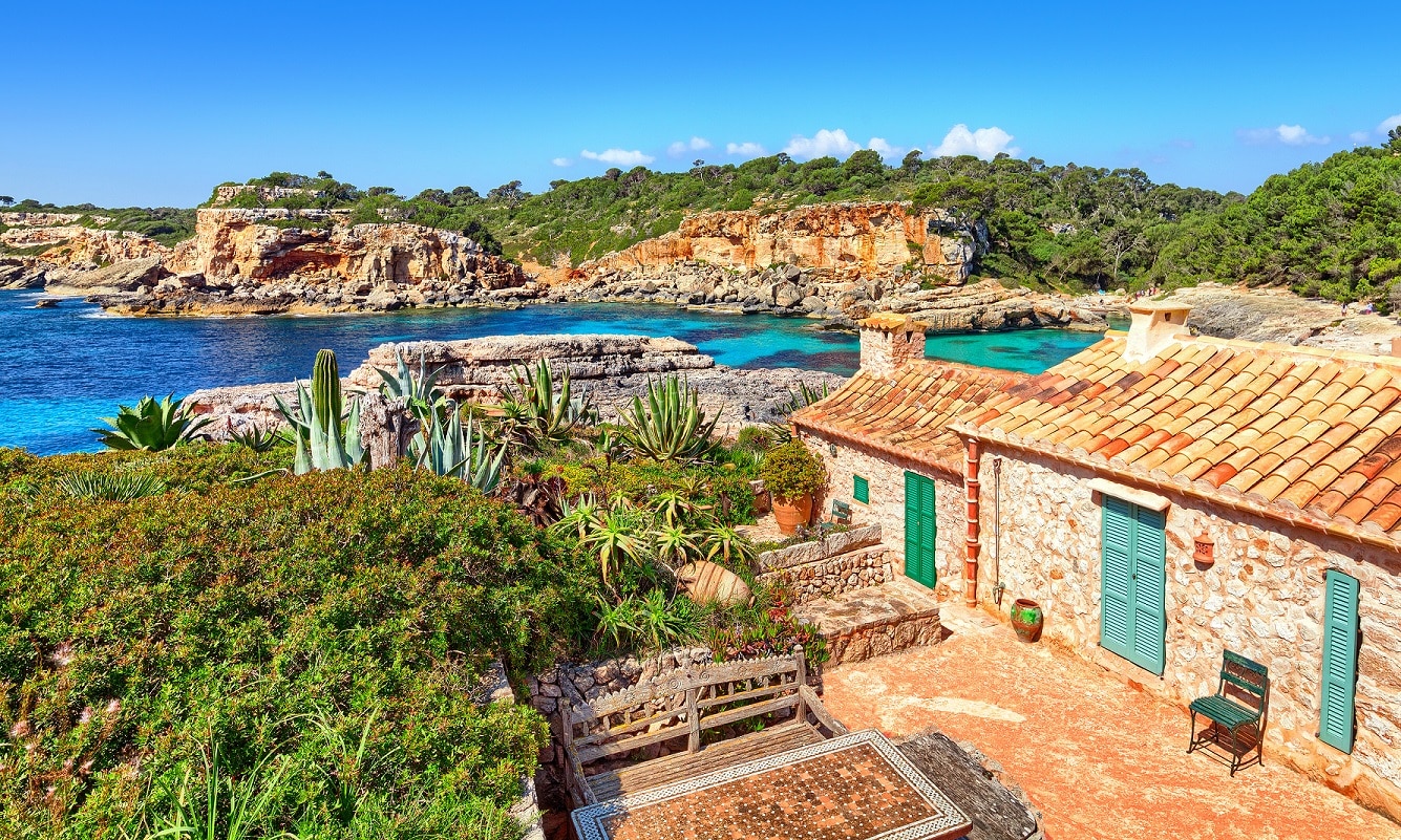 Mallorca Cala Almunia Spanien Landschaft mit Mittelmeer Traditionelle Finca im Vordergrund, im Hintergrund eine Mittelmeerbucht mit Felswänden und Wald.