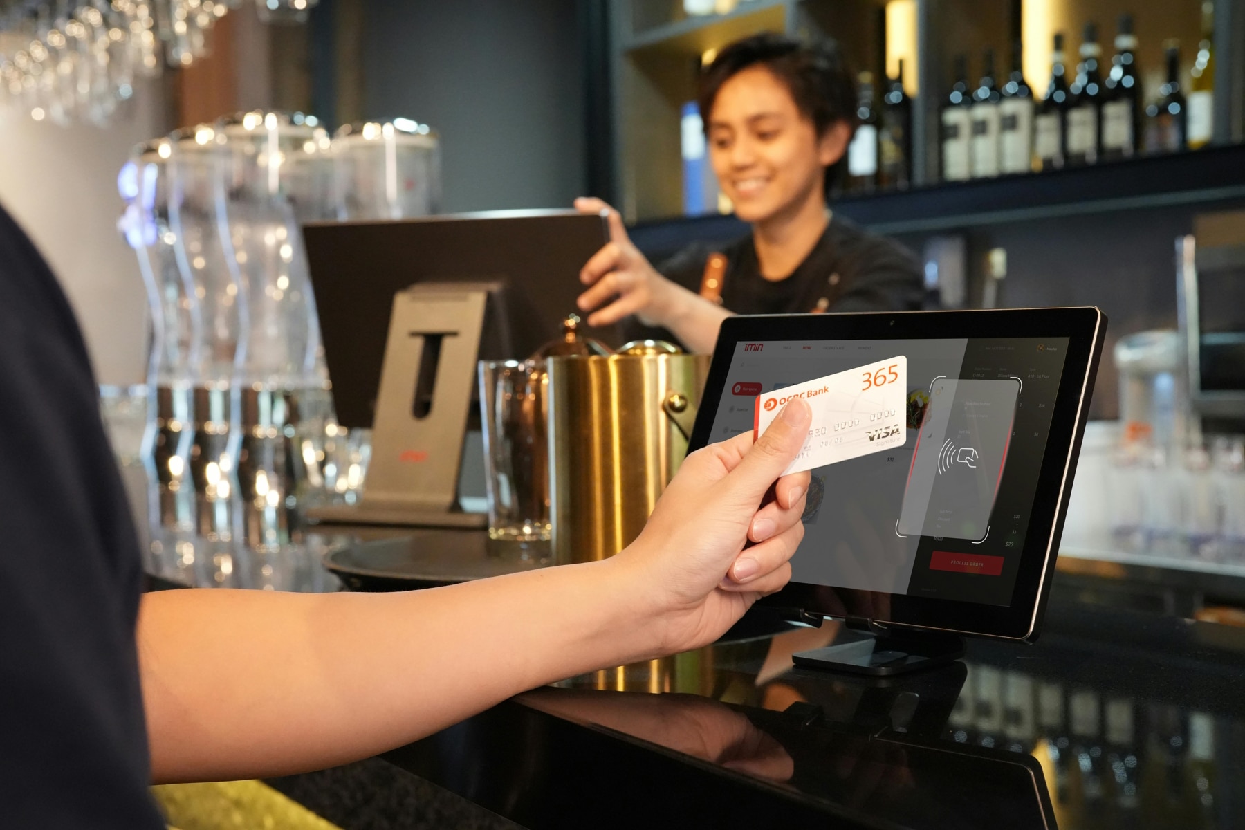 Bezahlen am Tresen Hand mit Kreditkarte bezahlt kontaktlos am Tresen eines Restaurants. Hinter der Bar ist eine Frau am Computermonitor.