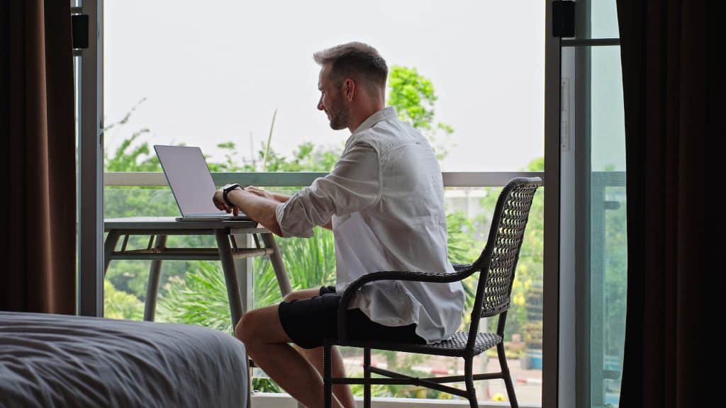 Remote Worker Typing On Laptop In Hotel Balcony Workspace Man Typing On Laptop At Hotel Balcony Workspace During Remote Work Routine, Digital Nomad Lifestyle And Flexible Online Business Concept