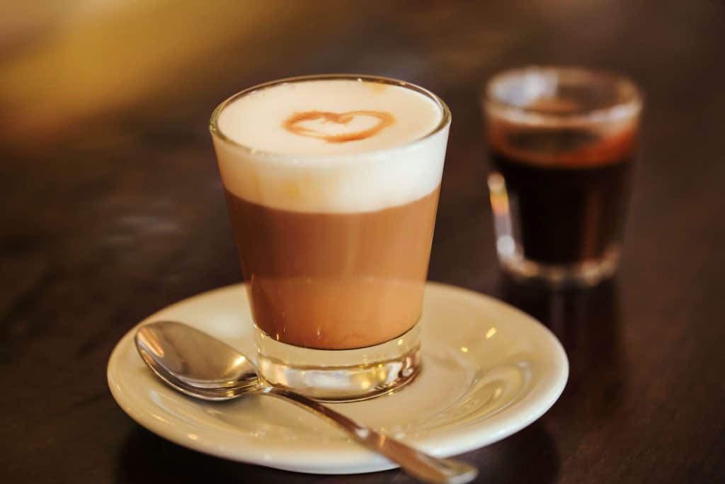 capuccino inside glass called „capo in b“ at Trieste Capucciono im Glas mit Herz im Milchschaum auf weißem Unterteller mit Löffel auf dunklem Holztisch, daneben im Hintergrundein Espresso im Glas