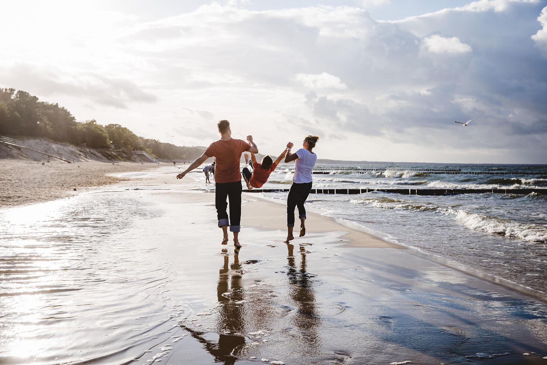 Vater, Mutter und Kind in der Mitte laufen am Ostseestrand entlang und schwingen das Kind an den Händen in die Luft.