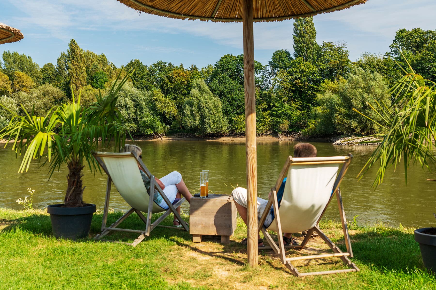 Urlaubsfeeling am Rhein Urlaubsfeeling am Rhein, 2 Liegestühle und ein Sonnenschirm aus Holz und Bamus am Rheinufer im Sommer mit 2 Personen, die ein großes Glas Bier auf einem Holztisch vor sich haben und aufs Wasser blicken