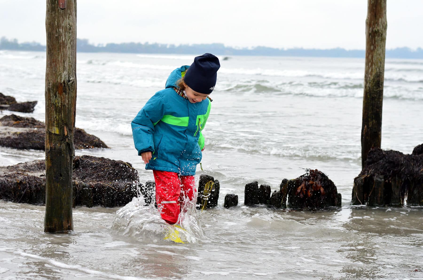 Kind planscht mit Gummistiefeln und Regenkleidung durch seichtes Wasser am Strand