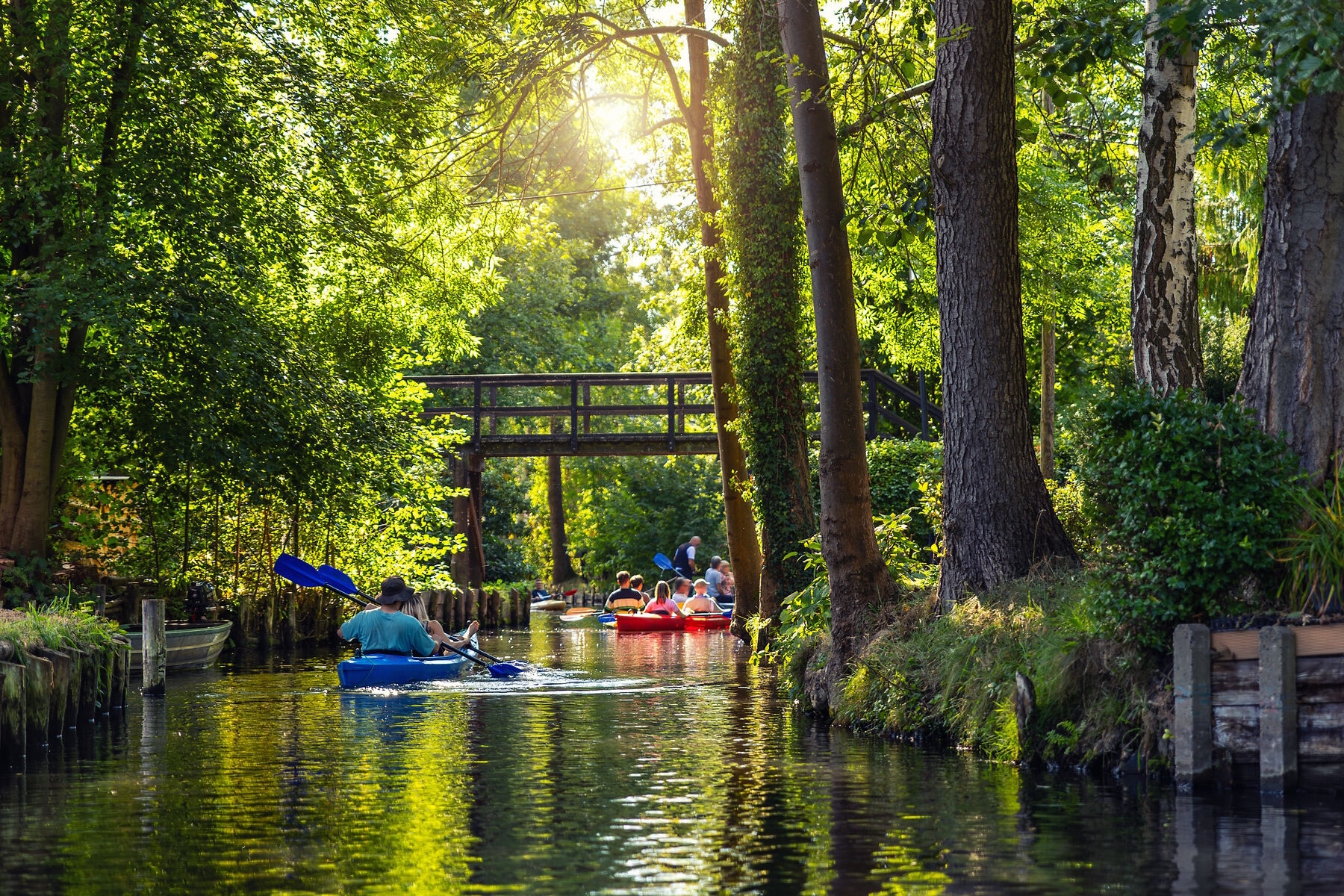 People paddling kayaks canal dense green forest bright summer sunlight reflections on water surface. Scenic wetland landscape creates peaceful atmosphere for outdoor recreation and travel exploration People paddling kayaks canal dense green forest bright summer sunlight reflections on water surface. Scenic wetland landscape creates peaceful atmosphere for outdoor recreation and travel exploration.