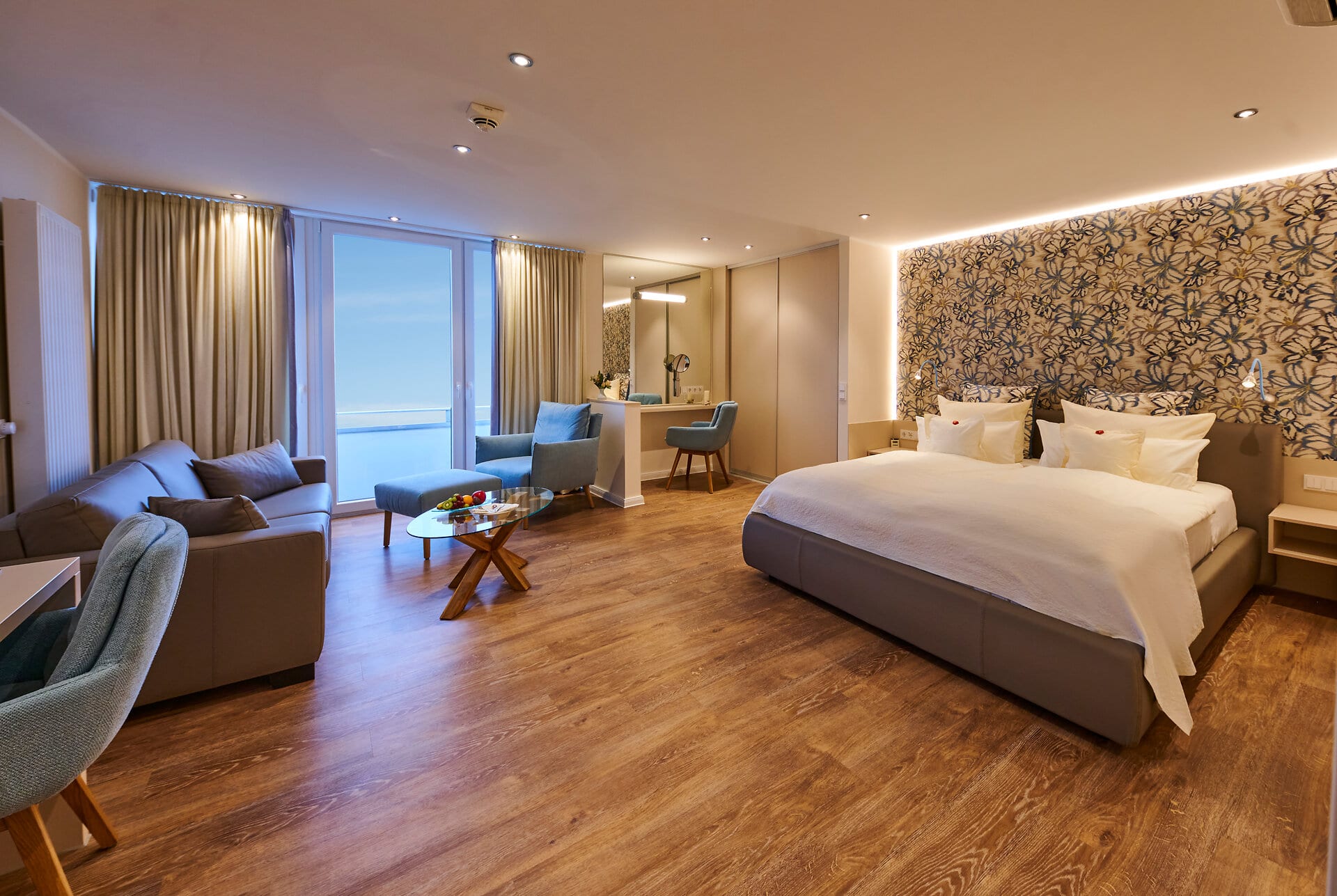 Suite Badhotel Sternhagen Großes Zimmer mit Doppelbett und Parkettboden, Loungeecke und bodentiefen Fenstern.