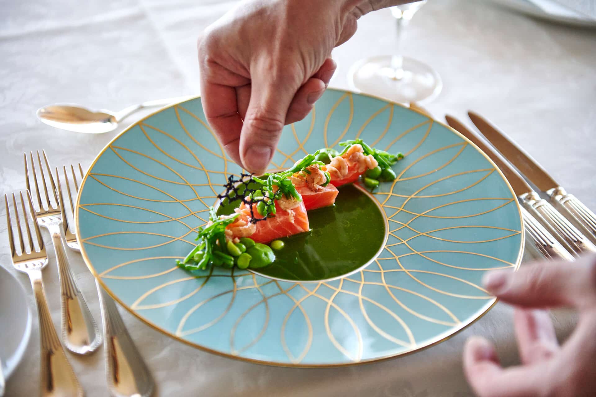 Foodbild Badhotel Sternhagen Nahaufnahme Hellblauer Teller mit goldenem Muster auf dem rosafarbener lachs mit grünem Gemüse und grüner Soße angerichtet ist. Eine Hand streut Kräuter über das Gericht